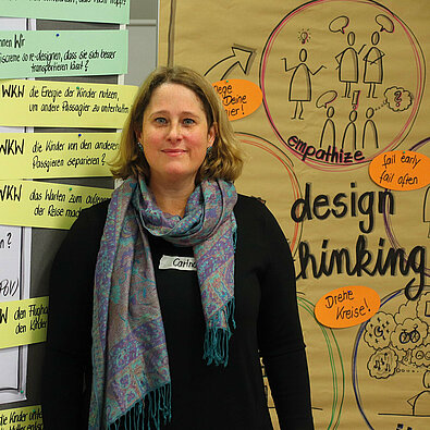 Referentin Carina Goffart steht neben Flipcharts o.ä. mit schwarzen Stichpunkten und diversen bunten Pappen, um das Design Thinking zu erklären