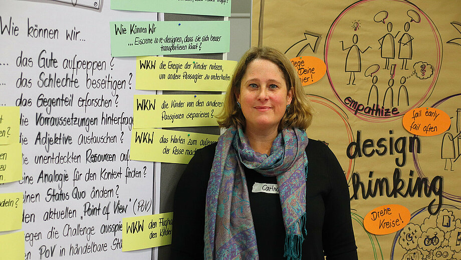 Referentin Carina Goffart steht neben Flipcharts o.ä. mit schwarzen Stichpunkten und diversen bunten Pappen, um das Design Thinking zu erklären