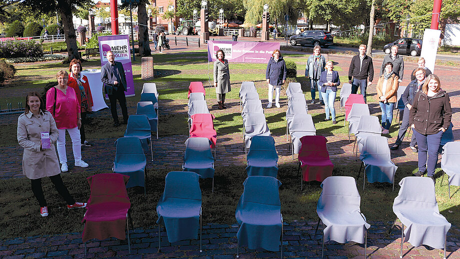 Bild mit mehreren Frauen die rechts und links neben grau und pink bedeckten Stühlen stehen. Die Farben sollen zeigen, dass der Anteil an Frauen in politischen Räten gering ist.