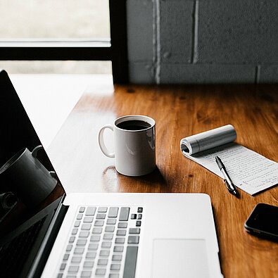 Laptop, Kaffeetasse, Notizblock, Stift und Handy auf einem Schreibtisch - Symbolbild