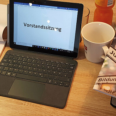 Laptop, auf dem der Bildschirm ein Dokument mit der großen Aufschrift Vorstandssitzung zeigt, daneben eine Kaffeetasse
